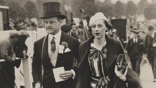 A black and white photo showing a gentleman in top hat and tail coat escorting a lady at a horse racing event